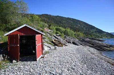 a redshed sitting on a rocky beach near the water at En perle i Trondheimsfjorden in Trondheim