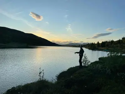 a man standing next to a lake holding a fishing rod at Cabin With A View And Close To Jotunheimen in Heggenes
