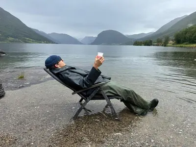 a man sitting in a chair next to a body of water at Living in a Nikolai Astrup motif 96m2 in Vassenden