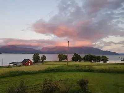 a red house in a field next to a body of water at Nicoll-huset in Ølve