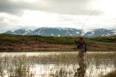 a man fishing in a river with mountains in the background at Geilolia Hyttetun in Geilo