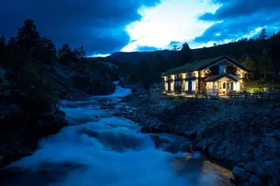 a cabin by a river at night with lights on at Billingen Seterpensjonat in Skjåk
